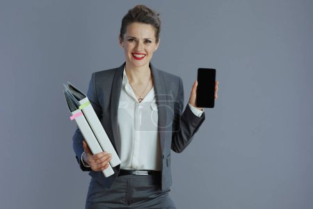 Foto de Sonriente mujer moderna de mediana edad propietaria de una pequeña empresa en traje gris con carpetas que muestran la pantalla en blanco del teléfono inteligente aislada sobre fondo gris. - Imagen libre de derechos