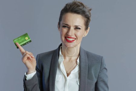 Foto de Sonriente mujer moderna de mediana edad trabajadora en traje gris con tarjeta de crédito aislada en gris. - Imagen libre de derechos