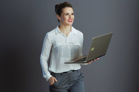 Foto de Sonriente joven mujer de negocios en blusa blanca con portátil contra fondo gris. - Imagen libre de derechos