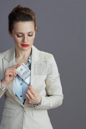 Photo for Trendy business woman in a light business suit hiding dollars money packs against gray background. - Royalty Free Image