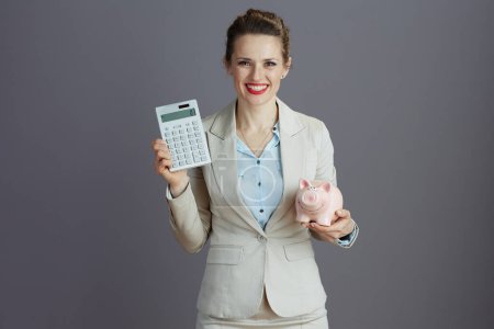 Photo for Happy modern small business owner woman in a light business suit with piggy bank and calculator isolated on grey background. - Royalty Free Image