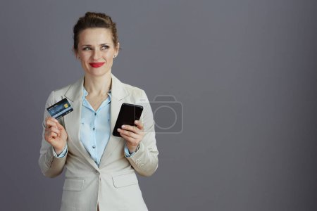 Photo for Smiling elegant female employee in a light business suit with smartphone and credit card isolated on gray. - Royalty Free Image