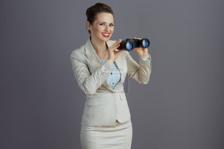 Téléchargez les photos : Souriant employé féminin à la mode dans un costume d'affaires léger avec jumelles isolées sur gris. - en image libre de droit