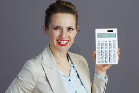 Photo for Smiling modern 40 years old woman employee in a light business suit with calculator against gray background. - Royalty Free Image