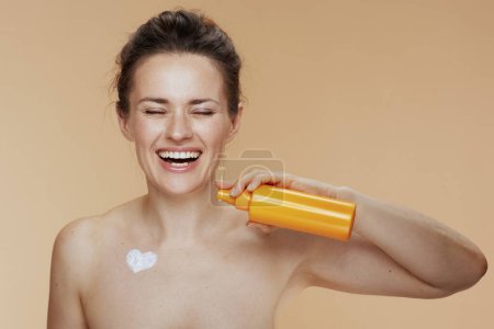Photo for Smiling modern 40 years old woman with cosmetic cream jar and heart shaped cream on chest isolated on beige background. - Royalty Free Image
