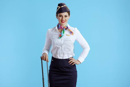 Foto de Sonriente elegante azafata mujer sobre fondo azul en uniforme con bolsa de carrito. - Imagen libre de derechos
