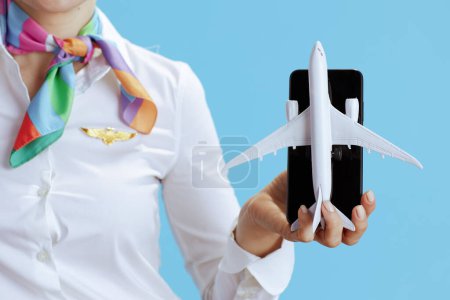 Foto de Primer plano de la azafata moderna sobre fondo azul en uniforme que muestra un poco de avión y teléfono inteligente. - Imagen libre de derechos