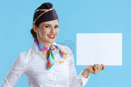 Téléchargez les photos : Heureuse hôtesse de l'air élégante femelle sur fond bleu en uniforme montrant feuille de papier blanc a4. - en image libre de droit