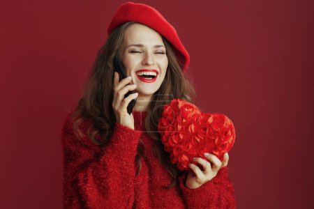 Téléchargez les photos : Joyeux Valentin. heureuse femme tendance en pull rouge et béret avec coeur rouge parlant sur un smartphone. - en image libre de droit