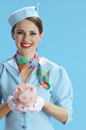 Photo for Smiling modern female stewardess on blue background in blue uniform with piggy bank. - Royalty Free Image