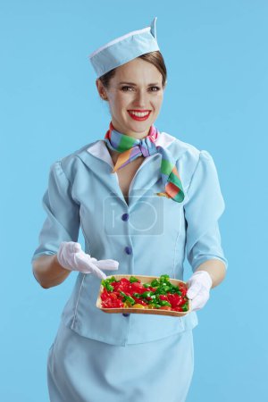 Photo for Smiling stylish flight attendant woman against blue background in blue uniform with candies. - Royalty Free Image
