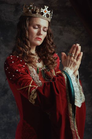 Photo for Medieval queen in red dress with crown praying on dark gray background. - Royalty Free Image
