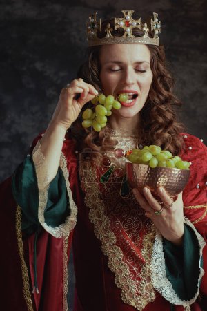 Foto de Reina medieval en vestido rojo con plato de uvas y corona sobre fondo gris oscuro. - Imagen libre de derechos