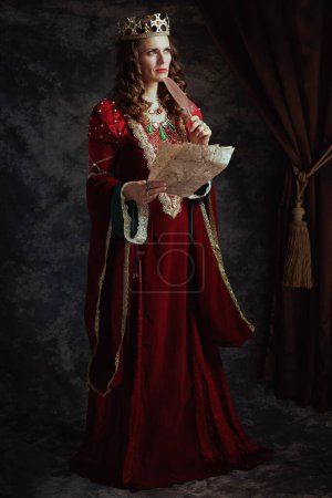 Foto de Retrato completo de la reina medieval pensativa en vestido rojo con pergamino y corona sobre fondo gris oscuro. - Imagen libre de derechos