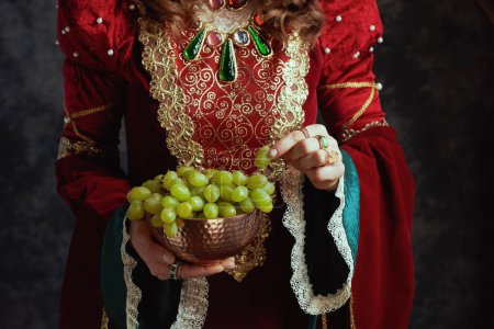 Téléchargez les photos : Gros plan sur la reine médiévale en robe rouge avec assiette de raisins sur fond gris foncé. - en image libre de droit