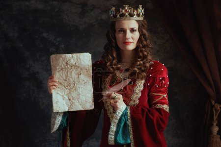 Photo for Smiling medieval queen in red dress with parchment and crown on dark gray background. - Royalty Free Image
