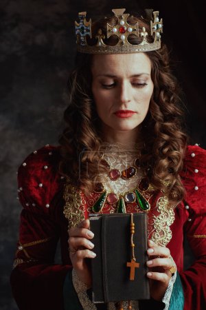 Photo for Medieval queen in red dress with book, rosary and crown on dark gray background. - Royalty Free Image