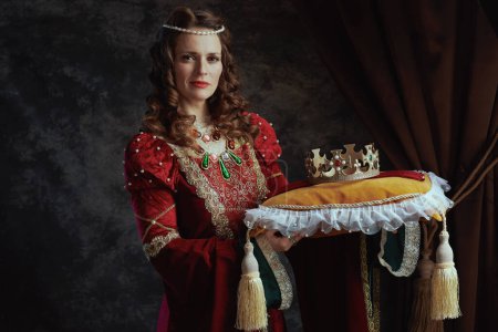Foto de Reina medieval en vestido rojo con corona sobre almohada sobre fondo gris oscuro. - Imagen libre de derechos