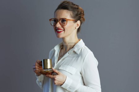 Foto de Empleada moderna de mediana edad feliz en blusa blanca con gafas y taza de café aislada sobre fondo gris. - Imagen libre de derechos