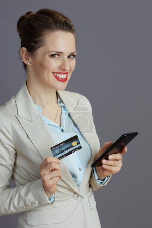 Foto de Sonriente mujer trabajadora elegante en un traje de negocios ligero con teléfono inteligente y tarjeta de crédito aislada sobre fondo gris. - Imagen libre de derechos