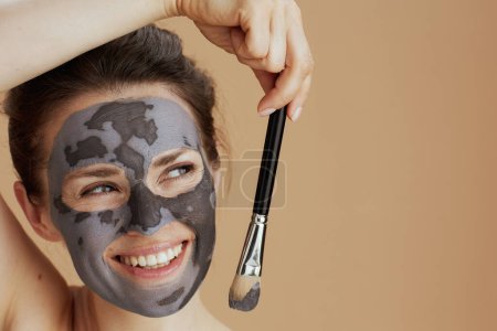 Téléchargez les photos : Femme souriante moderne de 40 ans avec brosse et masque cosmétique isolé sur fond beige. - en image libre de droit
