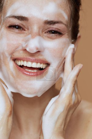 Foto de Sonriente hembra moderna con espuma facial limpiador lavado cara aislada sobre fondo beige. - Imagen libre de derechos