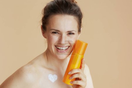 Téléchargez les photos : Femme souriante moderne de 40 ans avec pot de crème cosmétique et crème en forme de coeur sur la poitrine isolée sur fond beige. - en image libre de droit