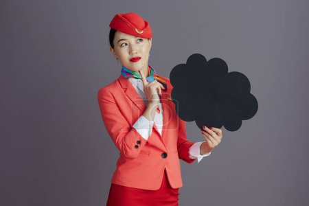 Foto de Pensativo moderno asiático femenino vuelo asistente en rojo falda chaqueta y sombrero uniforme mostrando en blanco nube forma tablero aislado en gris fondo. - Imagen libre de derechos
