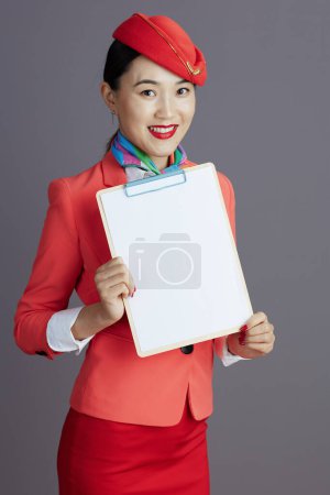 Foto de Feliz elegante asiática azafata de vuelo en falda roja, chaqueta y sombrero uniforme mostrando portapapeles en blanco sobre fondo gris. - Imagen libre de derechos