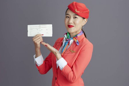 Téléchargez les photos : Heureuse hôtesse élégante asiatique femme en jupe rouge, veste et chapeau uniforme avec billets d'avion sur fond gris. - en image libre de droit