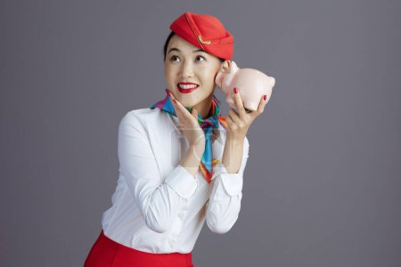 Photo for Cheerful modern asian female flight attendant in red skirt and hat uniform with piggy bank against gray background. - Royalty Free Image