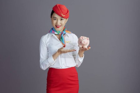 Photo for Smiling modern stewardess asian woman in red skirt and hat uniform with piggy bank isolated on grey background. - Royalty Free Image