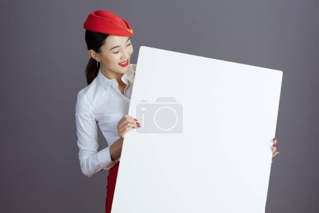 Photo for Smiling stylish asian female flight attendant in red skirt and hat uniform with blank billboard isolated on gray background. - Royalty Free Image