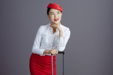 Foto de Pensativa elegante asiática azafata de vuelo en falda roja y sombrero uniforme con bolsa de viaje mirando espacio de copia sobre fondo gris. - Imagen libre de derechos