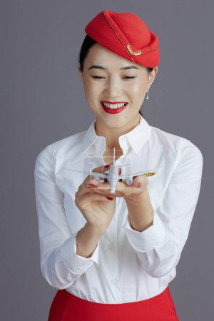 Photo for Smiling elegant asian female flight attendant in red skirt and hat uniform with a little airplane isolated on grey background. - Royalty Free Image