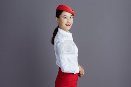 Photo for Smiling elegant asian female flight attendant in red skirt and hat uniform isolated on gray background. - Royalty Free Image