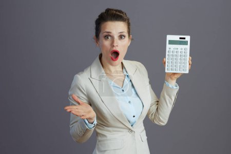 Photo for Shocked elegant middle aged woman employee in a light business suit with calculator isolated on grey. - Royalty Free Image