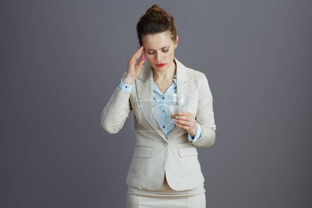 Foto de Acentuó la trabajadora moderna de 40 años en un traje de negocios ligero con vaso de agua y píldora aislada en gris. - Imagen libre de derechos