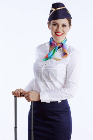Foto de Elegante azafata femenina sonriente sobre fondo blanco en uniforme con bolsa de ruedas. - Imagen libre de derechos