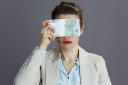 Photo for Trendy 40 years old business woman in a light business suit with euros money packs showing see no evil gesture isolated on gray background. - Royalty Free Image