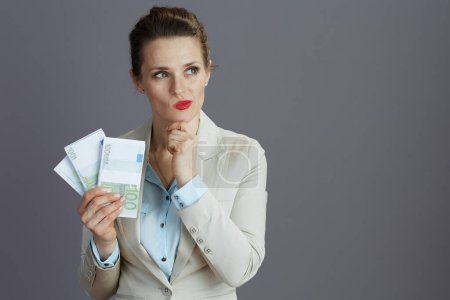 Foto de Empleada femenina elegante pensativa en un traje de negocios ligero con paquetes de dinero en euros contra el fondo gris. - Imagen libre de derechos