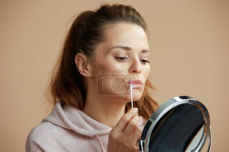 Téléchargez les photos : Femme avec herpès sur les lèvres et miroir appliquant pommade en utilisant coton-tige sur fond beige. - en image libre de droit