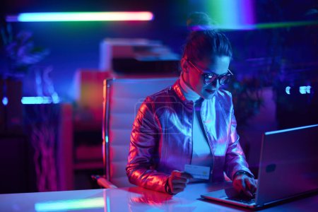 Téléchargez les photos : Neon metaverse concept futuriste. femme d'affaires moderne dans des lunettes avec ordinateur portable et carte de crédit dans le bureau moderne. - en image libre de droit