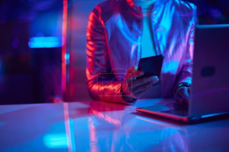 Photo for Neon metaverse futuristic concept. Closeup on woman with laptop using smartphone applications in office. - Royalty Free Image