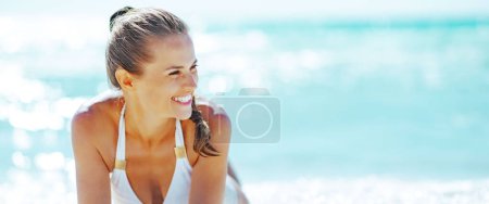 Téléchargez les photos : Jeune femme souriante assise au bord de la mer et regardant au loin - en image libre de droit