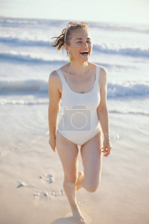 Téléchargez les photos : Portrait pleine longueur d'une femme élégante heureuse en maillot de bain blanc à la plage marchant. - en image libre de droit