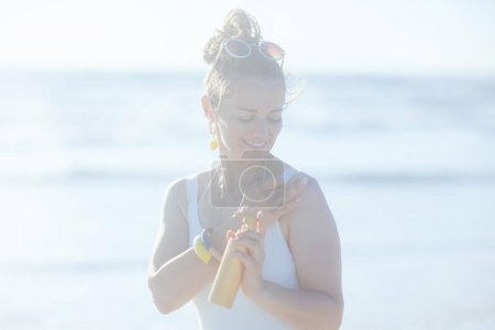 Téléchargez les photos : Heureux moderne femelle en maillot de bain blanc à la plage à l'aide de crème solaire. - en image libre de droit