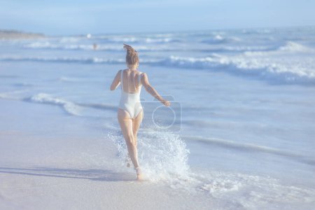 Téléchargez les photos : Vu de derrière moderne 40 ans femme en maillot de bain blanc à la plage avoir du plaisir. - en image libre de droit