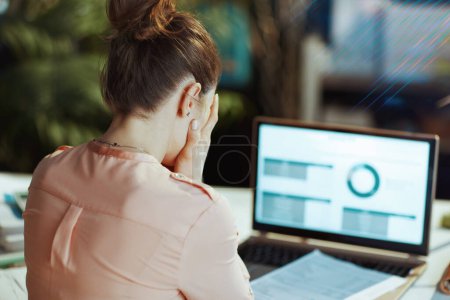 Photo for Seen from behind modern woman with documents and laptop having headache in modern office. - Royalty Free Image