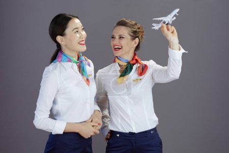 Photo for Smiling stylish flight attendant women in blue skirt, white shirt and scarf with a little airplane isolated on gray background. - Royalty Free Image